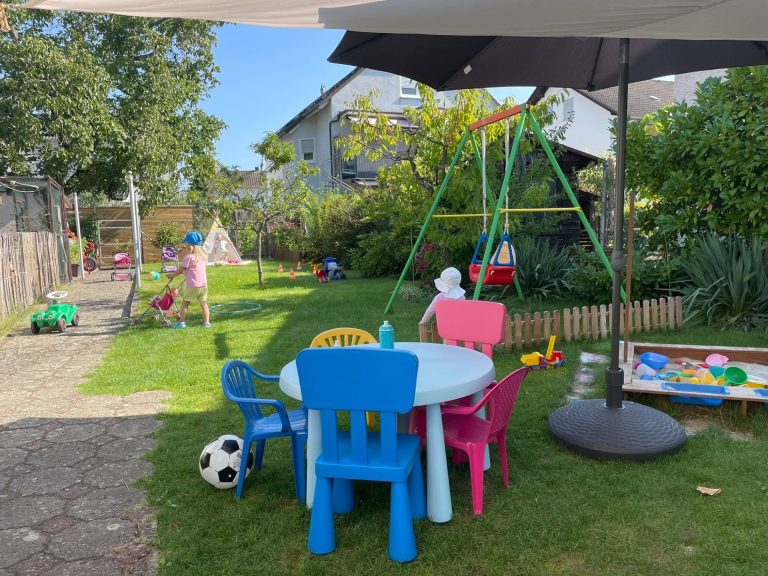Buntes Spielbereich im Garten mit kleinen Möbeln und einem Kind an einem Tisch.